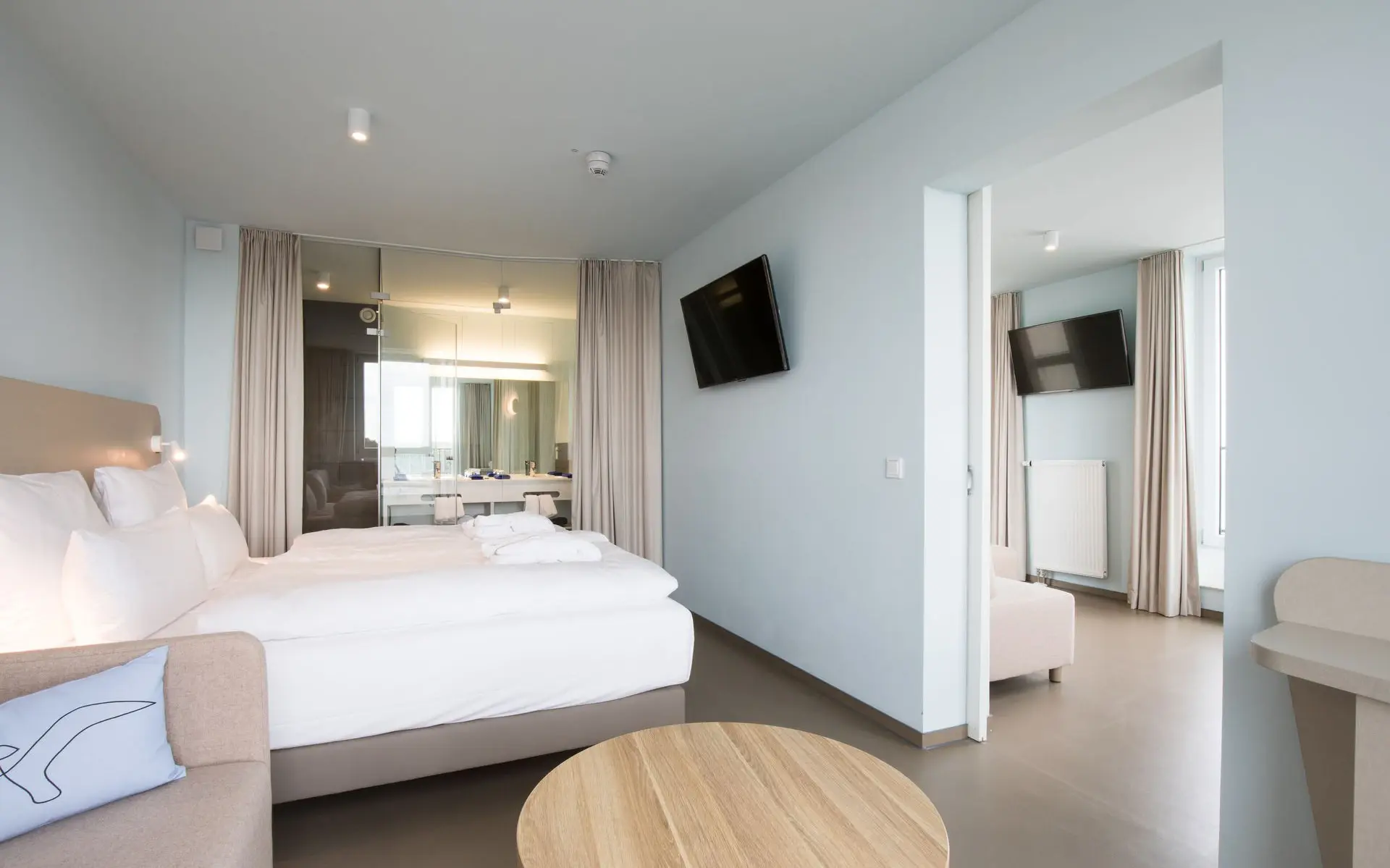Ein helles Hotelzimmer mit einem kleinen Sofa mit Couchtisch und einem Bett dahinter. Im Hintergund befindet sich ein Badezimmer mit Glaswand, welche durch einen Vorhang abgetrennt werden kann. Durch eine offene Tür ist ein Wohnbereich mit Sofa und Fernseher zu sehen.