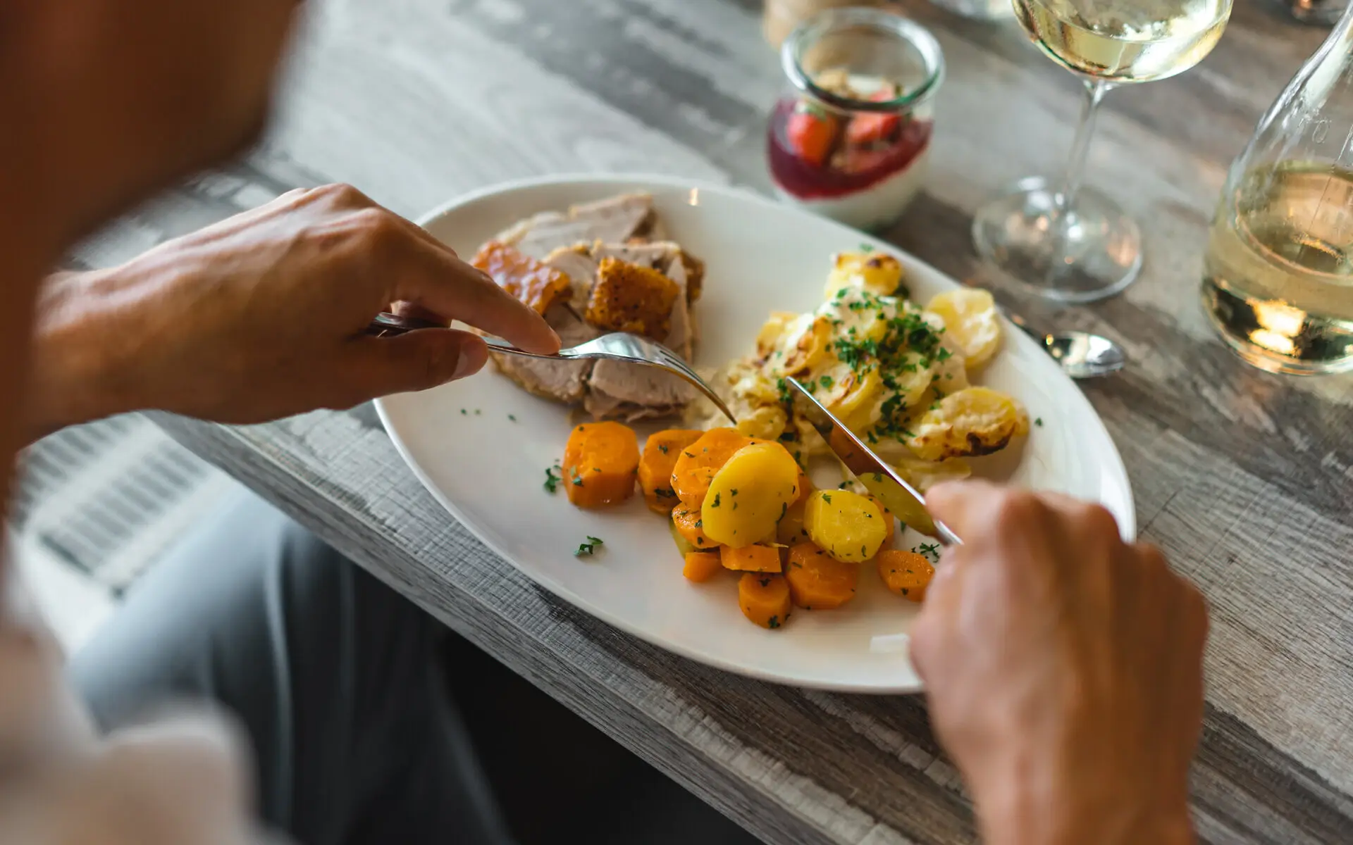 Abendessen aja Warnemünde Eine Person sitzt an einem Tisch und hat einen Teller mit gekochten Karotten, Kartoffelgratin und Fisch vor sich, das er mit einem Messer und einer Gabel isst.