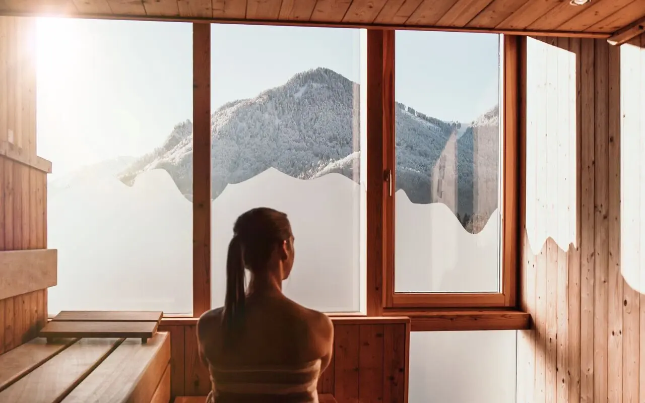 Eine Frau sitzt in einer Holzsauna mit Blick auf die schneebedeckten Berge und entspannt.