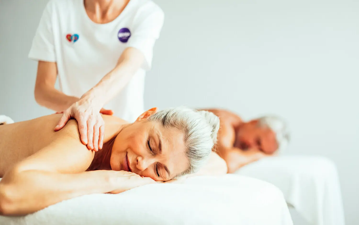 Anwendungsraum NIVEA Haus Travemünde Eine Frau liegt rückenfrei und mit einem Dutt entspannt auf einer Massageliege und wird von einer Frau in weißem Oberteil massiert. Hinter ihr liegt ein Mann, ebenfalls rückenfrei, auf einer weiteren Liege und entspannt.