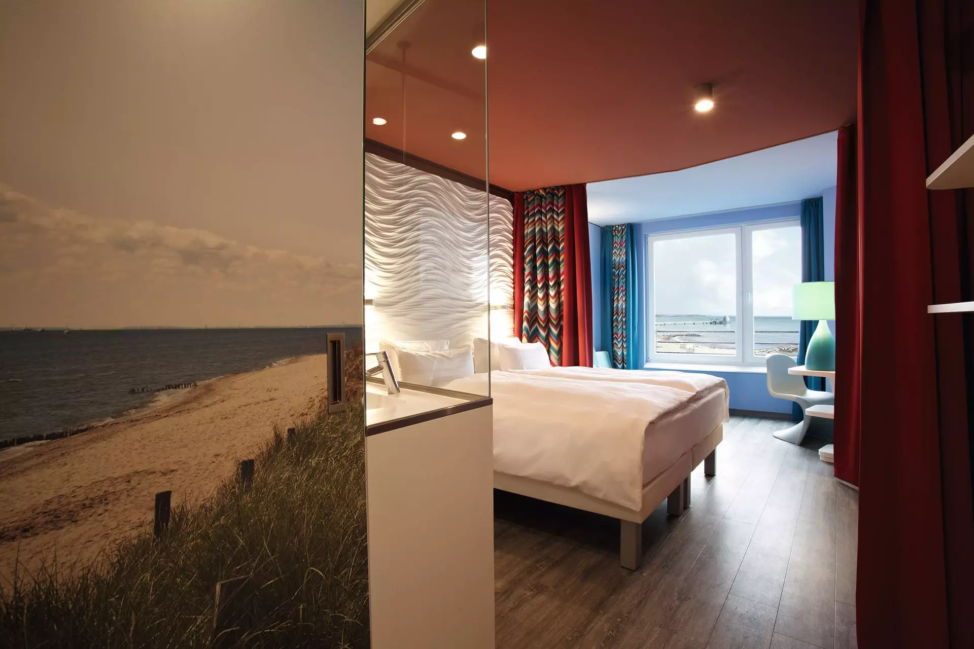 Ein gemütlich beleuchtetes Hotelzimmer. Die Tapete im Flur zeigt einen Strandzugang, dahinter liegt das Badezimmer mit Glaswand und ein Bett mit weißgewelltem Kopfteil. Ein zurückgezogener Vorhang trennt den dahinter liegenden Wohnbereich mit kleinem Sofa und Schreibtisch neben einem großen Fenster vom Schlafbereich.