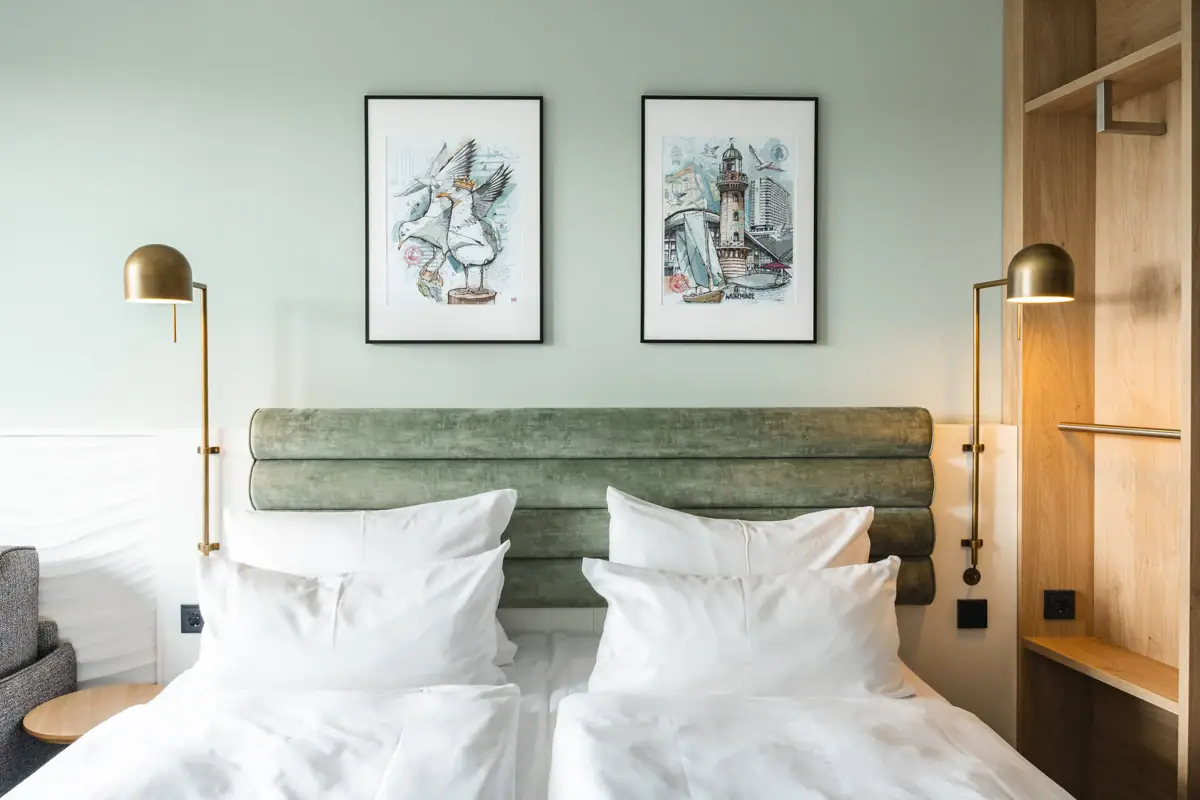 Neues Doppelzimmer im aja Warnemünde Bett mit weißen Kissen und grünem Kopfteil, flankiert von zwei Lampen.