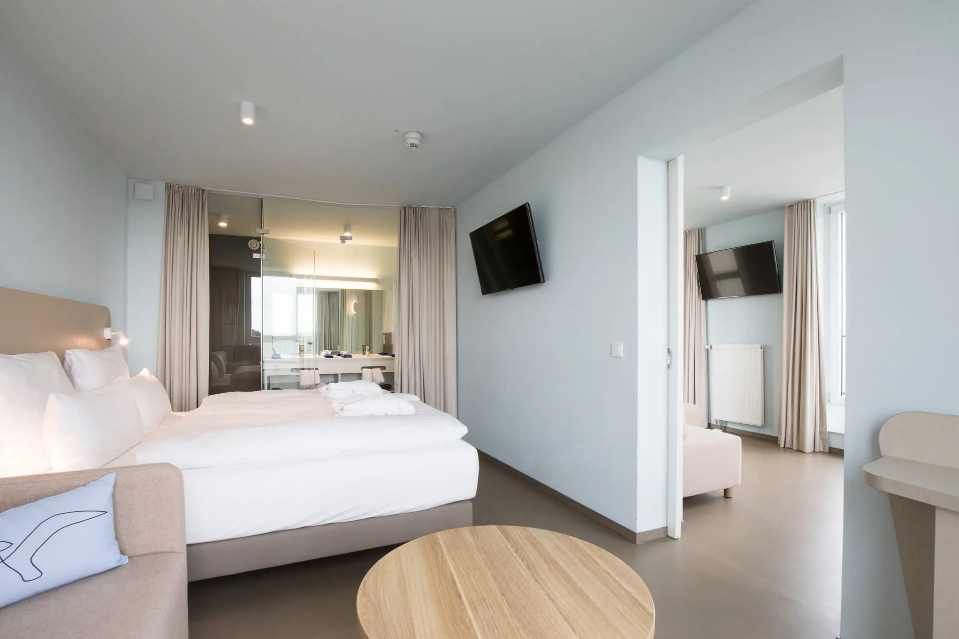 Suite im aja Travemünde Ein Zimmer mit einem Bett und einem Tisch, im Hintergrund ein zweites Zimmer, getrennt durch eine Tür.