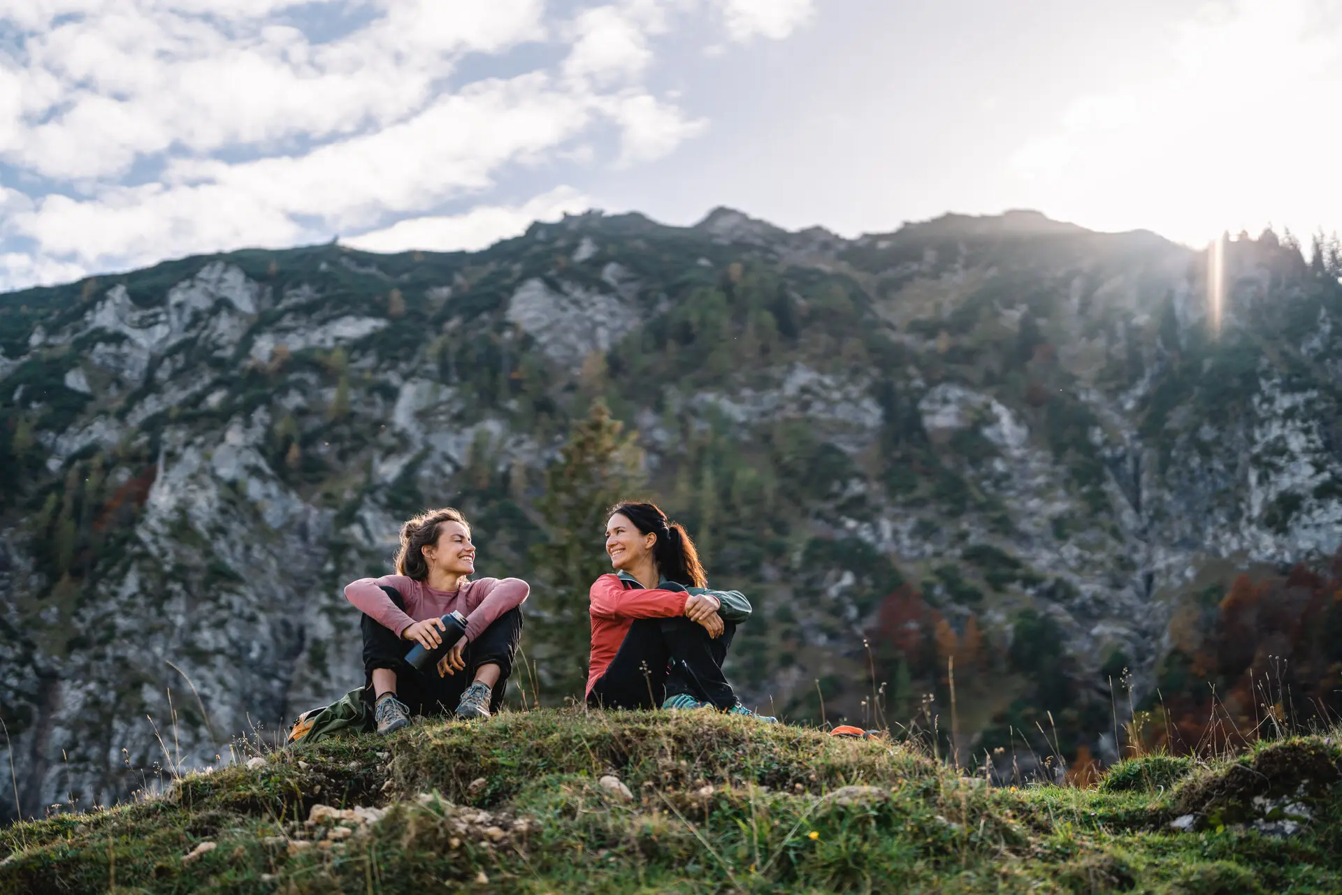 Zwei Frauen sitzen auf einem Hügel beim Wandern, mit Bergen im Hintergrund.