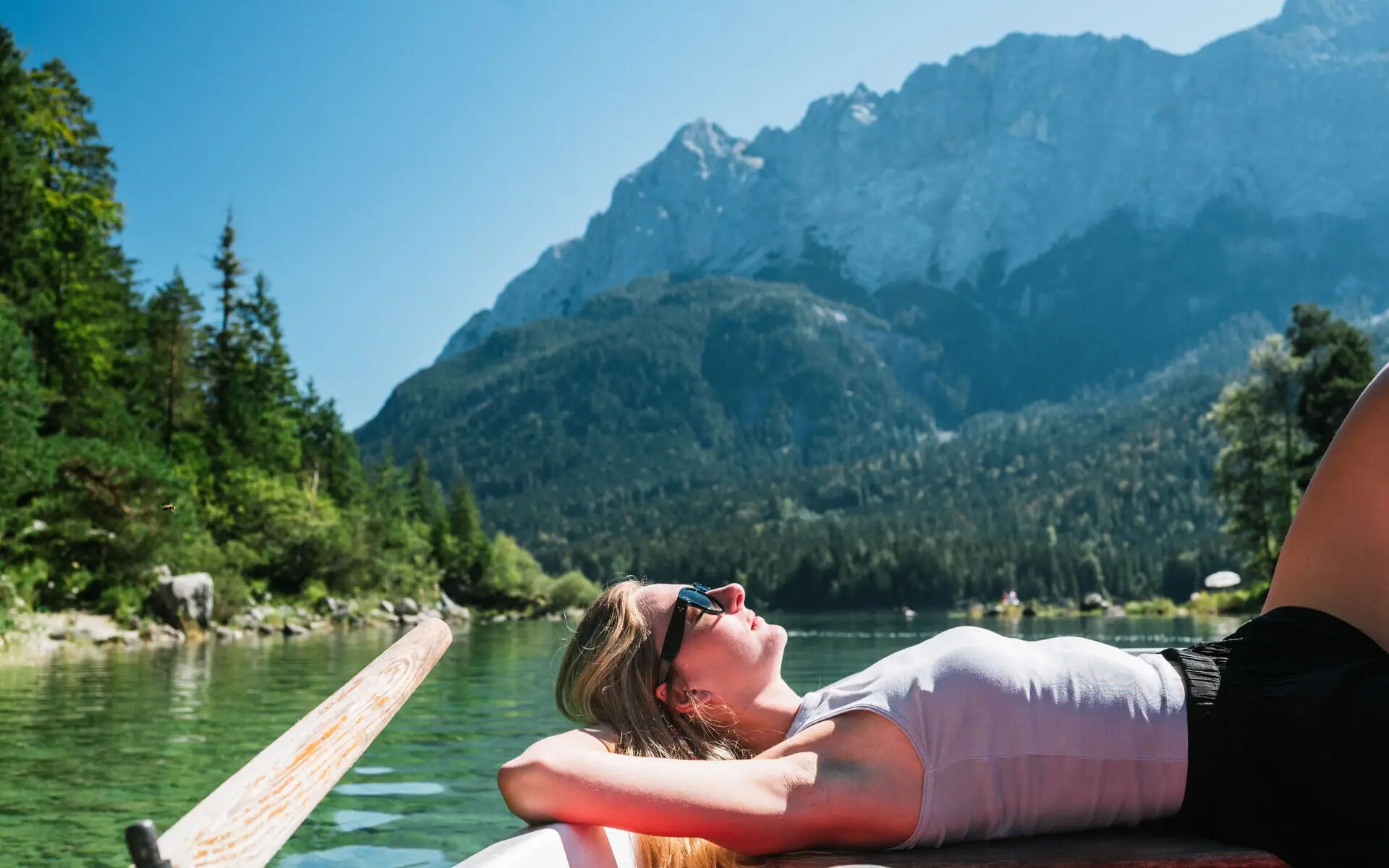 Sommerurlaub Eibsee Eine Frau liegt entspannt auf einem Boot am Eibsee. Die warme Sonne scheint ihr in das Gesicht und im Hintergrund erstreckt sich eine grüne Landschaft aus Bergen und Wäldern.