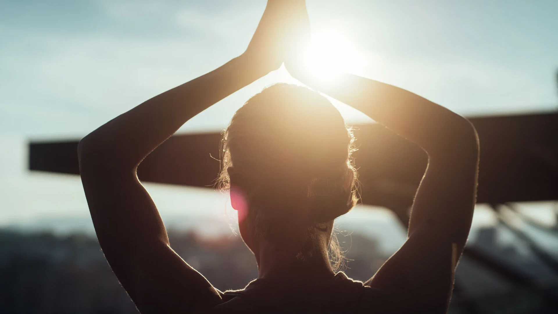 Eine Frau steht auf einer Terrasse und macht eine Yogaübung. Die aufgehende Sonne strahlt am Himmel.