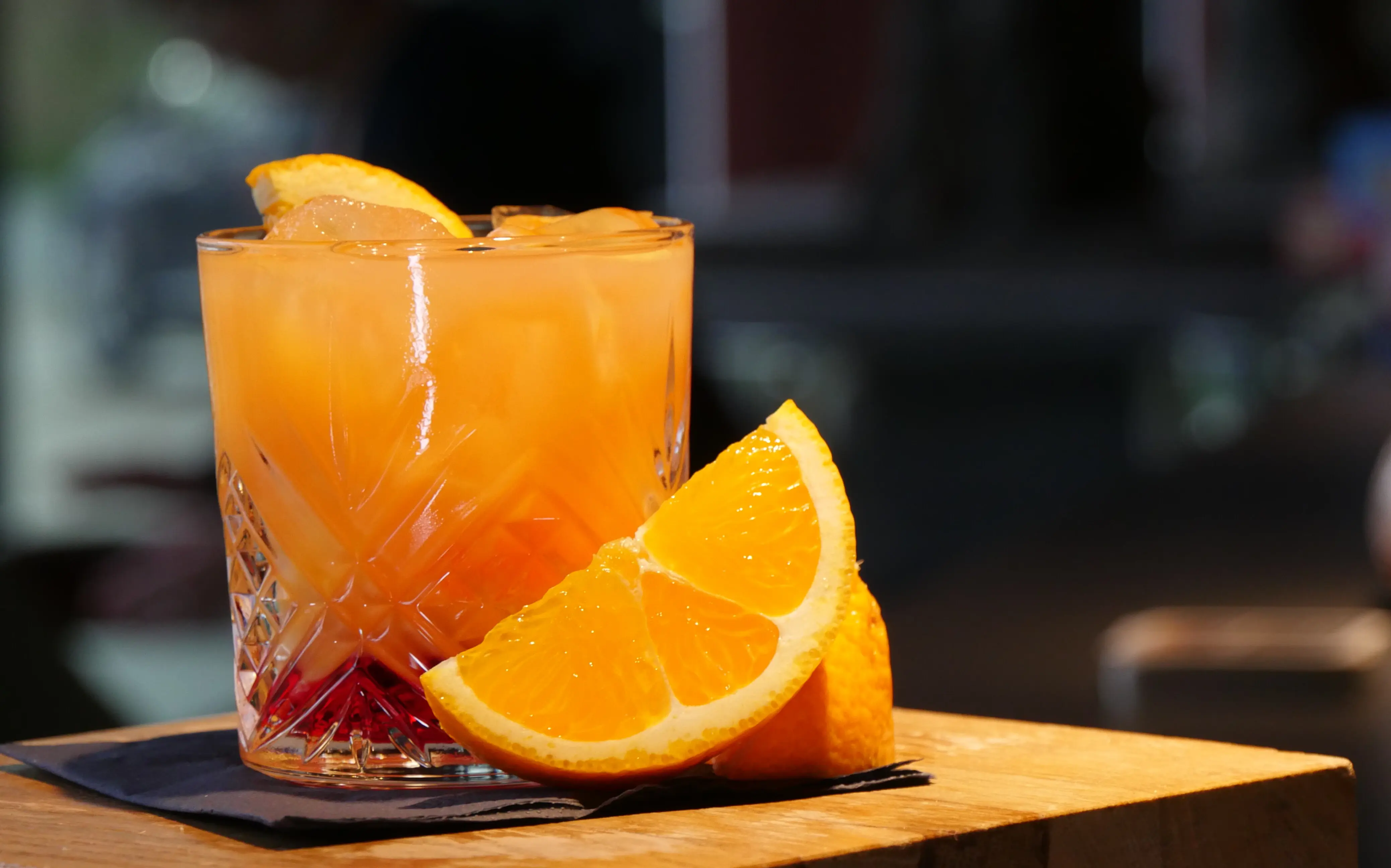 Drink Ein orangefarbenes Getränk mit Eiswürfeln und Orangenscheiben steht auf einer schwarzen Servierte auf einem kleinen Holztisch.