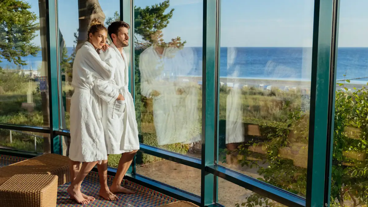 Wellnessbereich Ein Mann und eine Frau in Bademänteln stehen in einem Raum mit großen Fenstern.