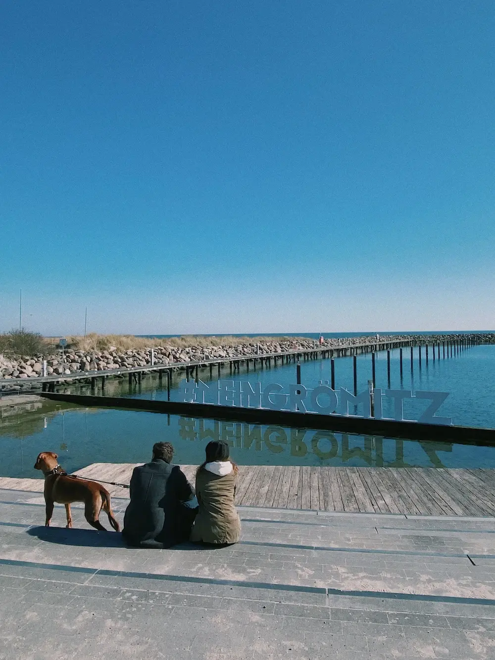Grömitzurlaub mit Hund Menschen sitzen mit einem Hund auf einem Dock und schauen aufs Wasser.