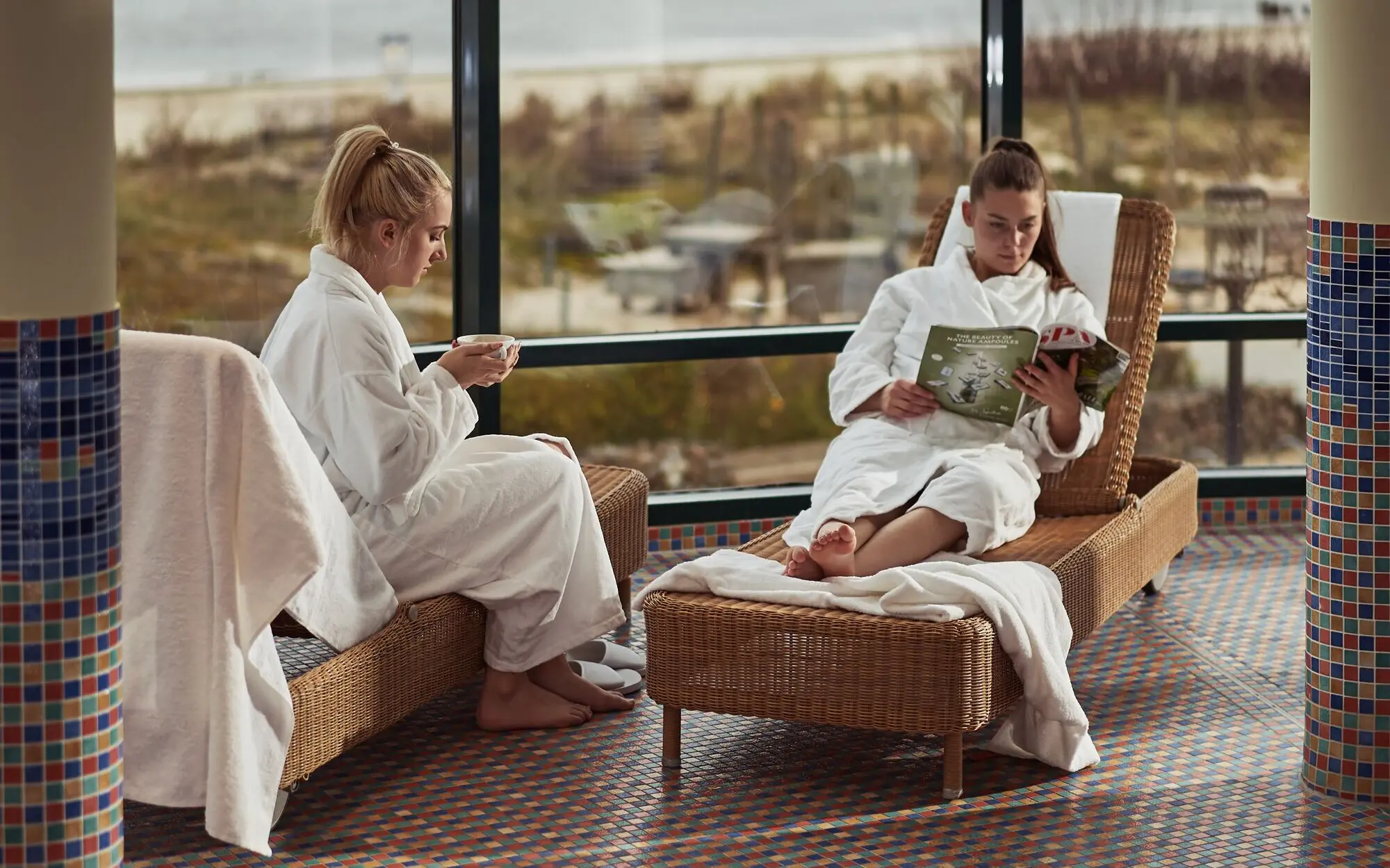 Wellness aja Bansin Zwei Frauen entspannen sich in einem Spa auf zwei Liegen mit Blick auf das Meer.Die eine hält eine Tasse in den Händen und die andere liest eine Zeitschrift.