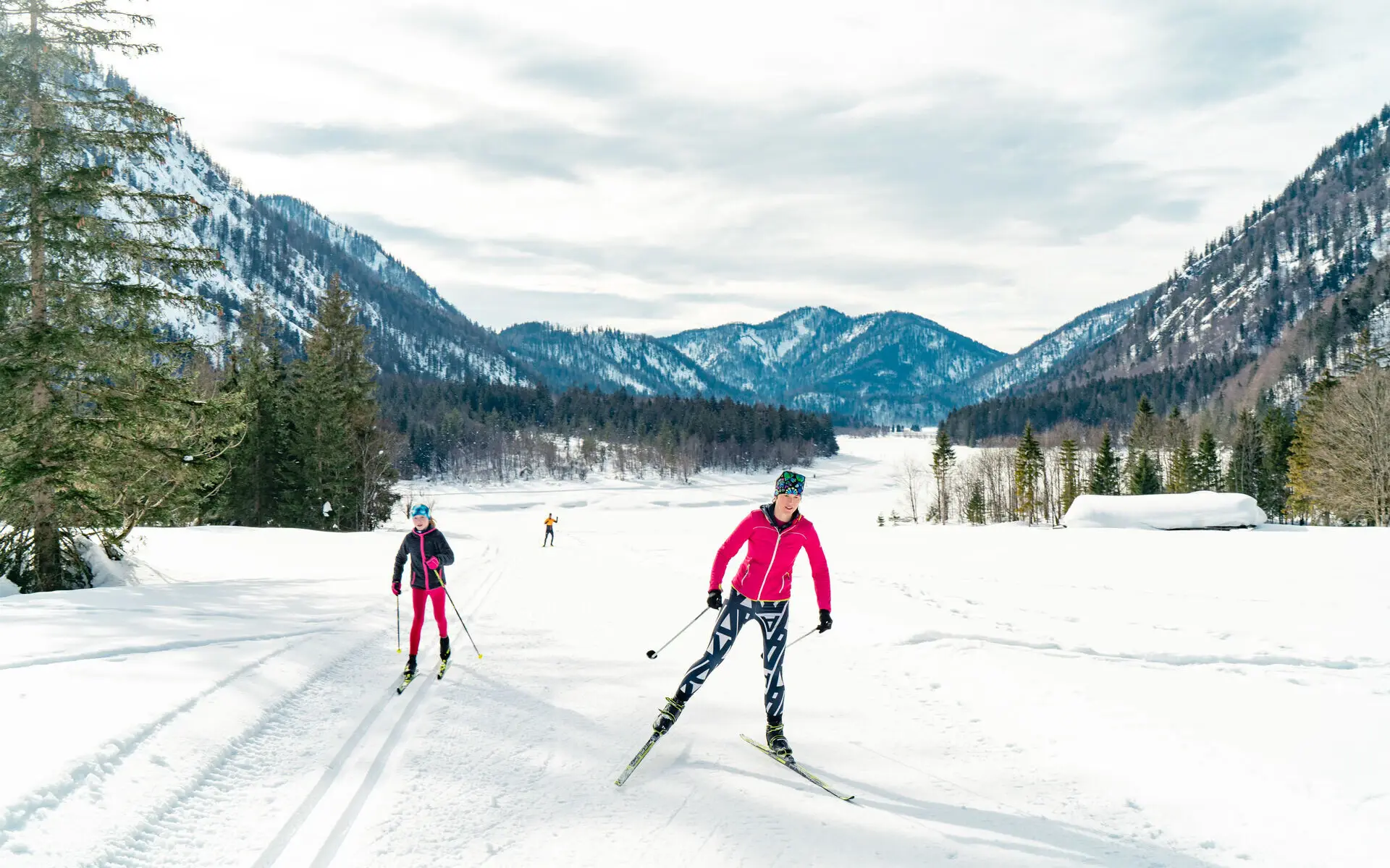 Zwei Frauen auf Skiern fahren durch den Schnee zwischen Bergen und Wäldern entlang.