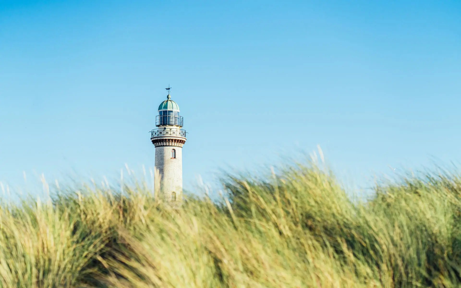 Zwischen im Wind wehenden Dünen steht in der Ferne ein heller Leuchtturm an einem sonnigen Tag.