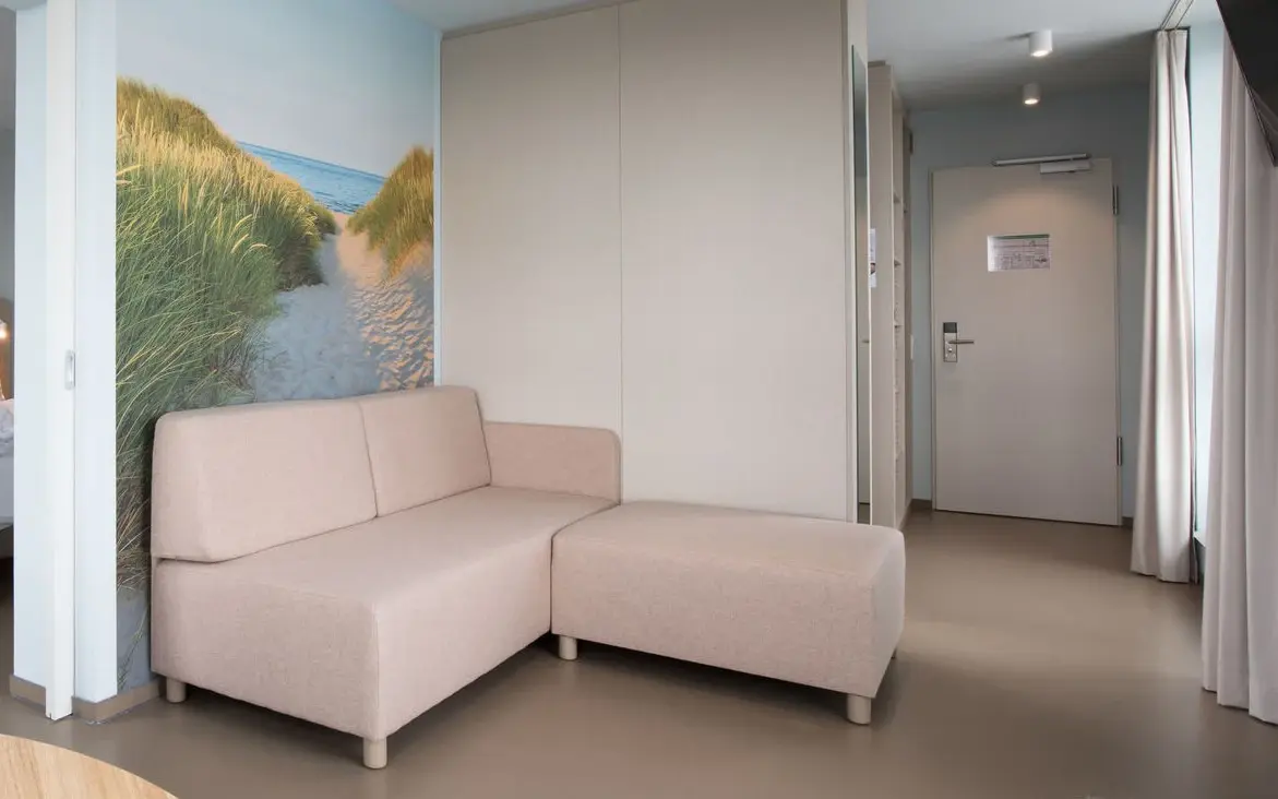 Ein helles Wohnzimmer mit einem beigen Sofa hinter dem ein großes Bild mit Strandzugang hängt.
