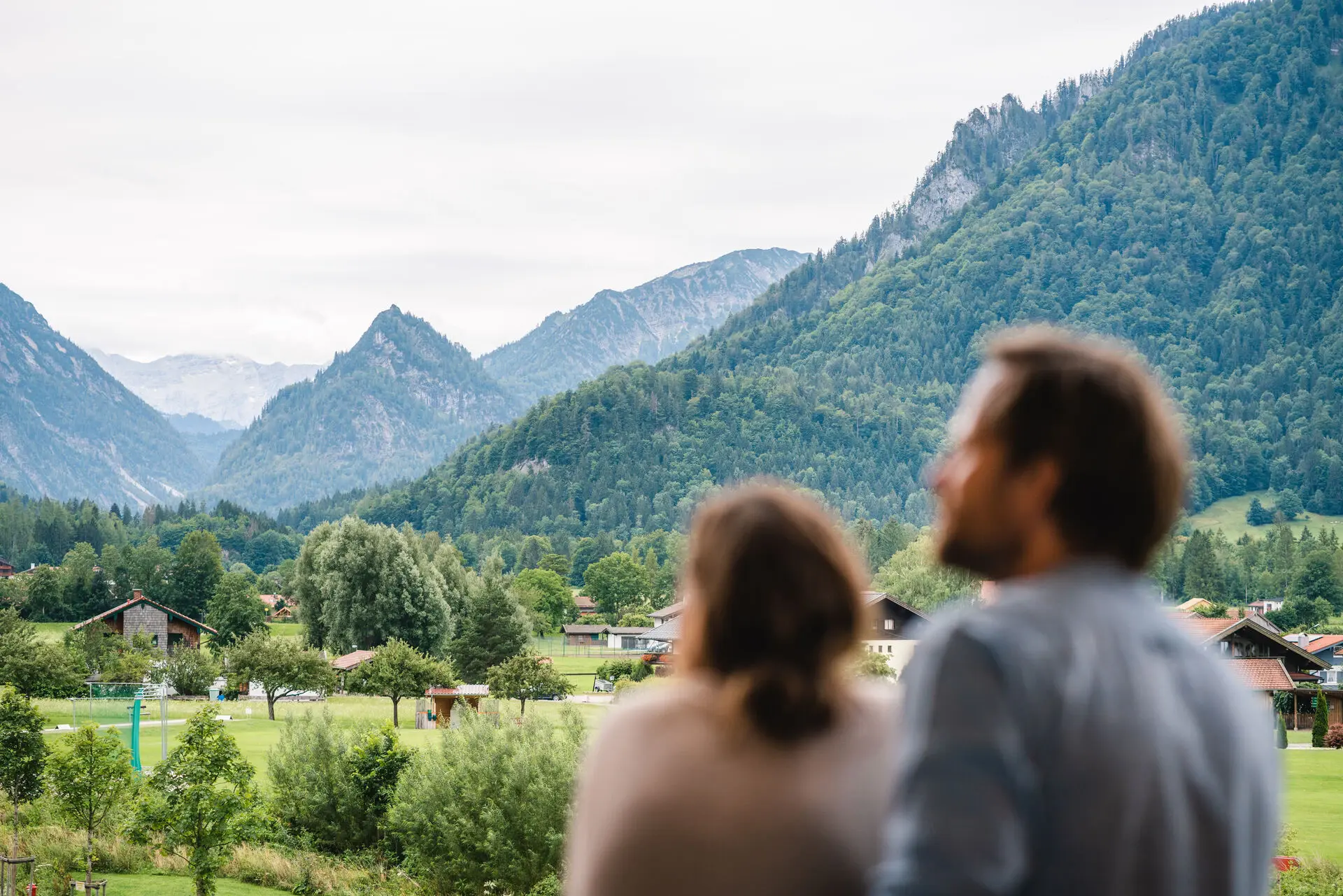 Ein Paar steht auf einem Balkon und schaut auf die umliegende Berg- Waldkulisse.