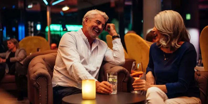 Ein älteres Paar sitzt entspannt in einer stilvollen Lounge bei gedämpfter Beleuchtung. Beide lachen herzlich, während sie ihre Getränke genießen. Ein älteres Paar sitzt entspannt in einer stilvollen Lounge bei gedämpfter Beleuchtung. Beide lachen herzlich, während sie ihre Getränke genießen.