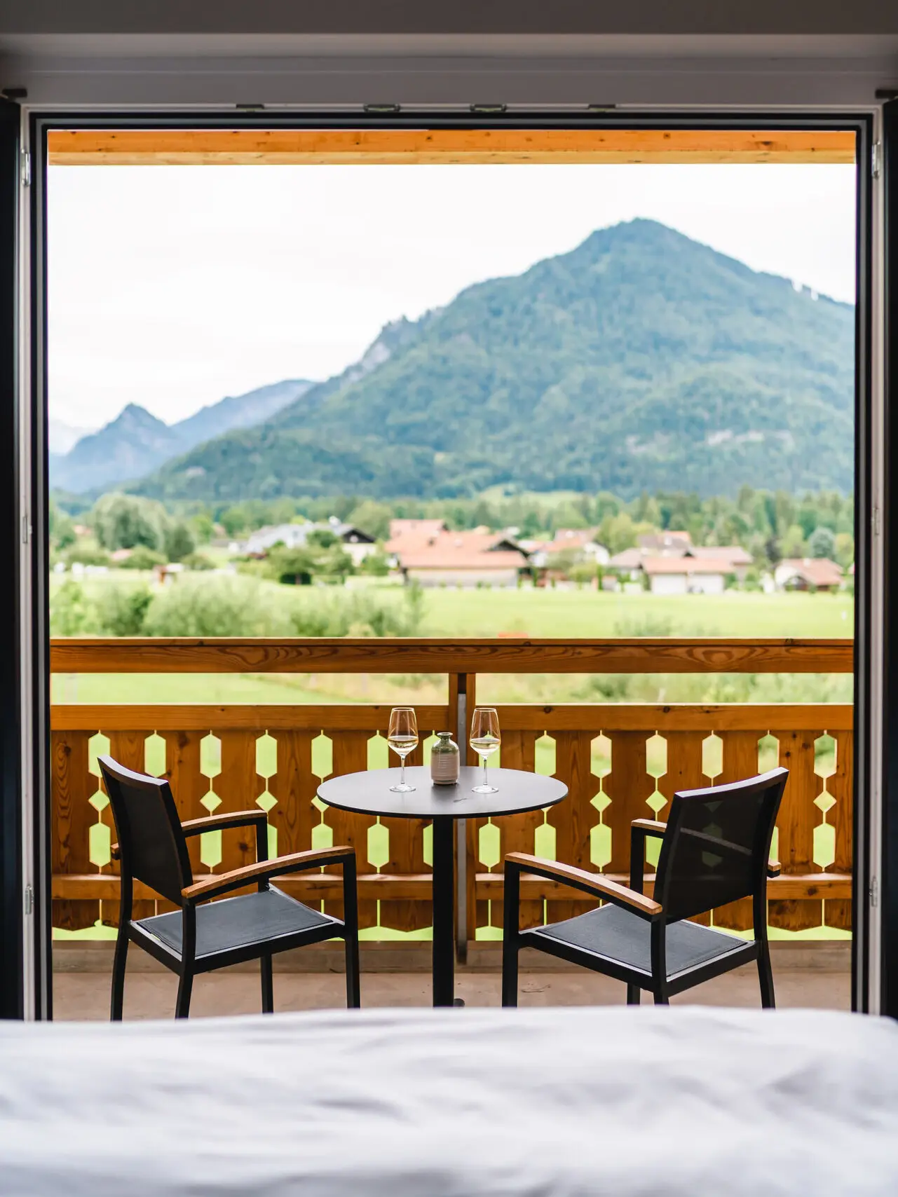 Auf einem Balkon mit Holzgeländer stehen zwei Stühle an einem kleinen runden Tisch mit Weingläsern. Im Hintergrund erstrecken sich grüne Wiesen und Berge.